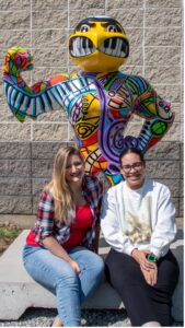 Rachel Hansen (left) and Sadé Head (right) sit in front of "The Musical World of Imagination" Herky.
